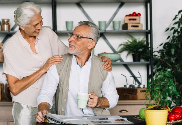 Smiling elderly couple in a kitchen, one holding a mug and newspaper, surrounded by plants and fresh produce.