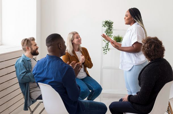 A woman stands speaking to a small group of people sitting in chairs, who are listening and clapping.