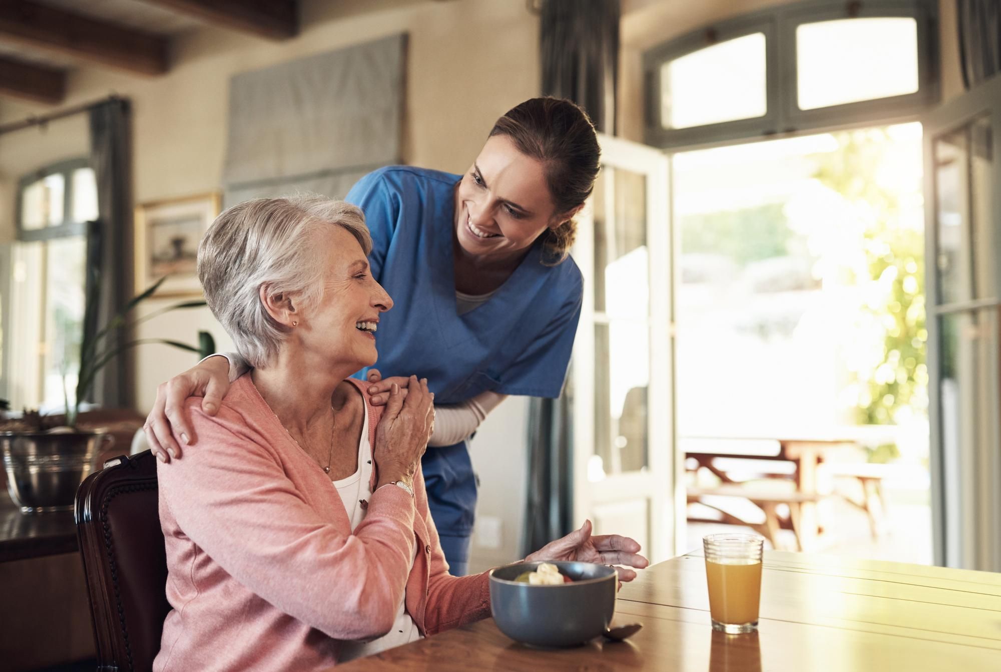 Caregiver in blue scrubs smiling at a seated elderly woman at a table with a bowl and a glass of juice.