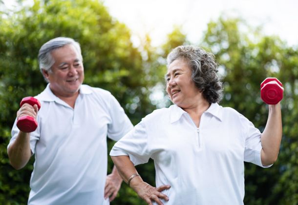 Smiling seniors in white shirts exercise outdoors with red dumbbells, enjoying a bright, green background.