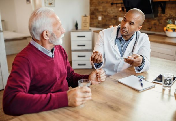 A doctor explains medication to an older man at a kitchen table; both hold glasses of water.