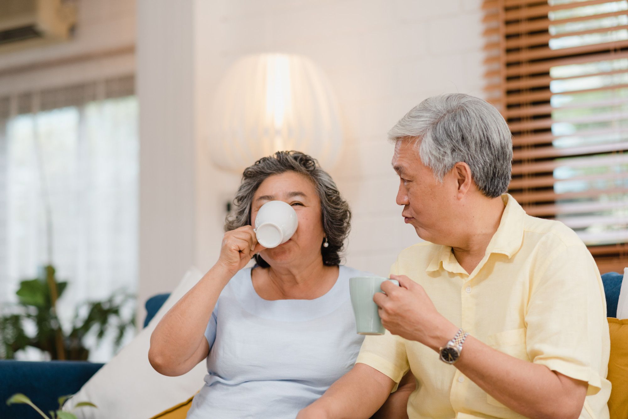 Older couple sitting on a couch at home, enjoying hot drinks and smiling together.