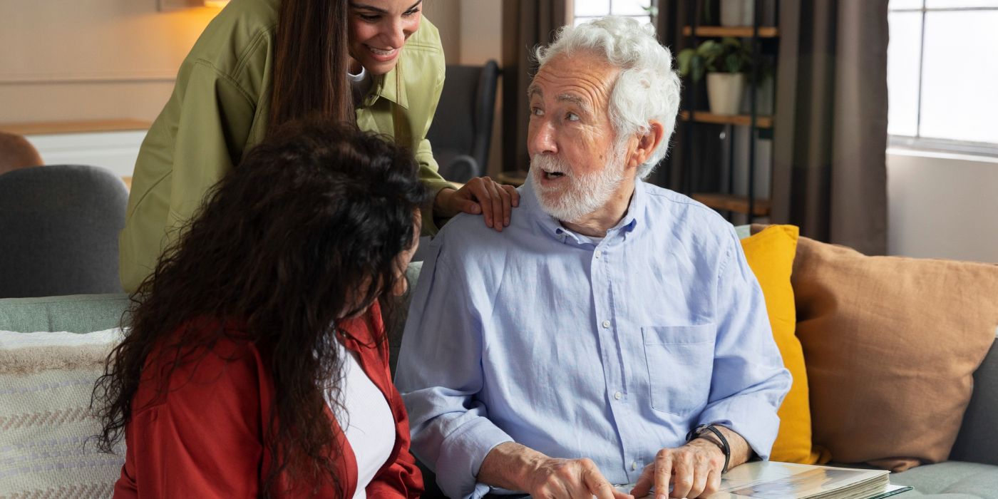 Older man showing a photo album to two women on a couch, all smiling and engaged in conversation.