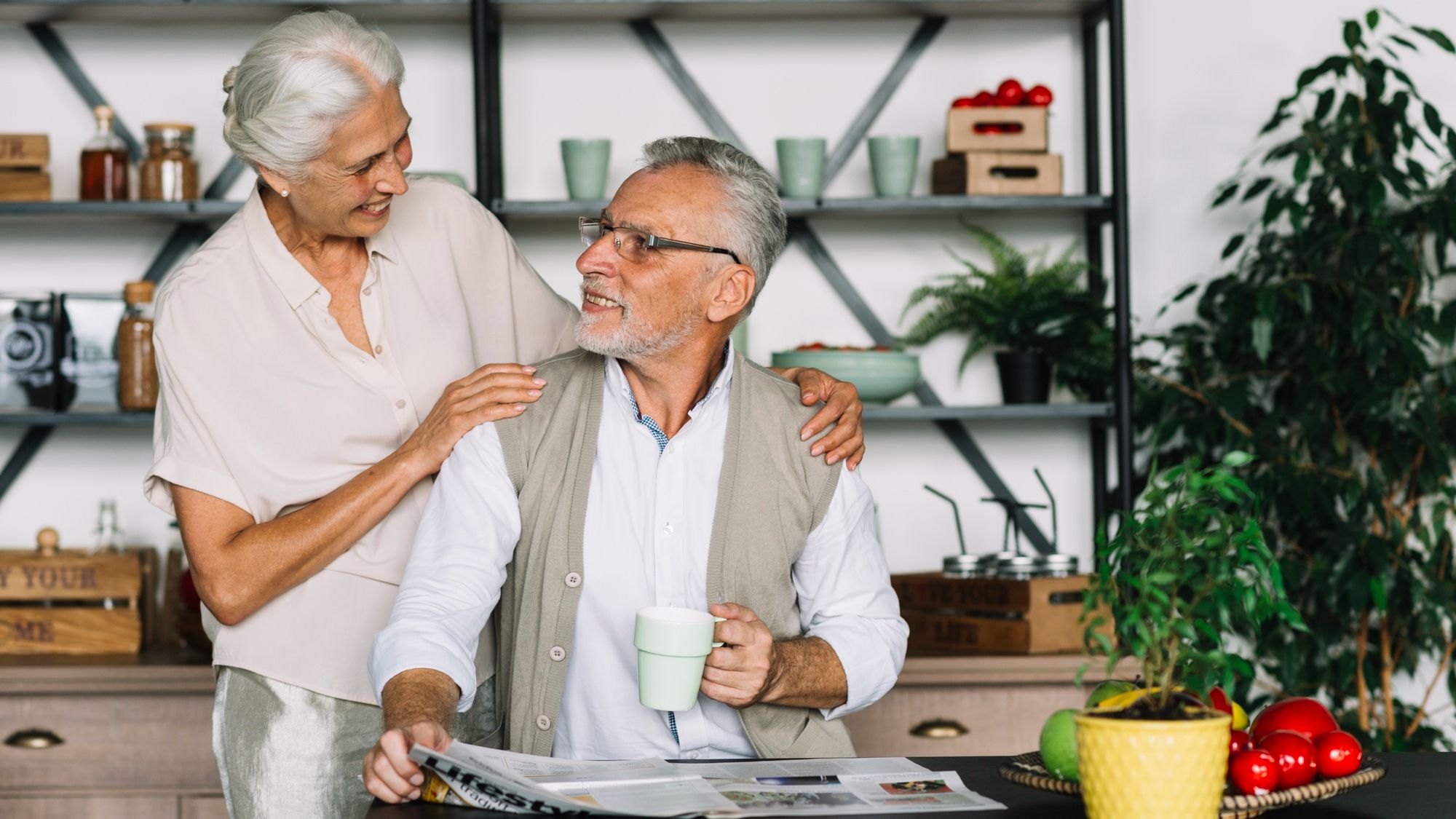 Smiling elderly couple in a kitchen, one holding a mug and newspaper, surrounded by plants and fresh produce.