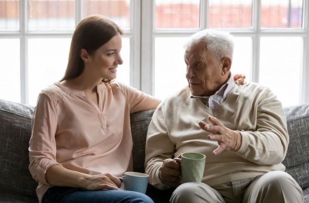 A young woman and an elderly man sitting on a sofa talking while holding mugs.