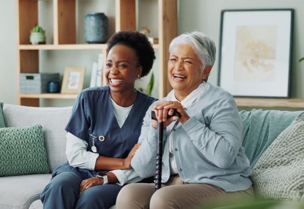 A caregiver and an older woman sitting on a sofa, both laughing.