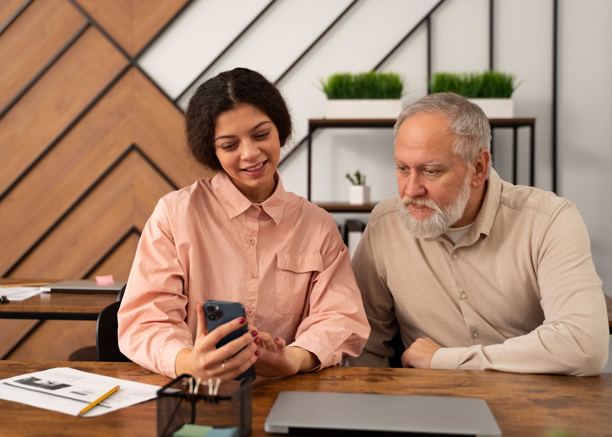 A young woman shows something on her phone to an older man as they sit together at a desk in a modern office.