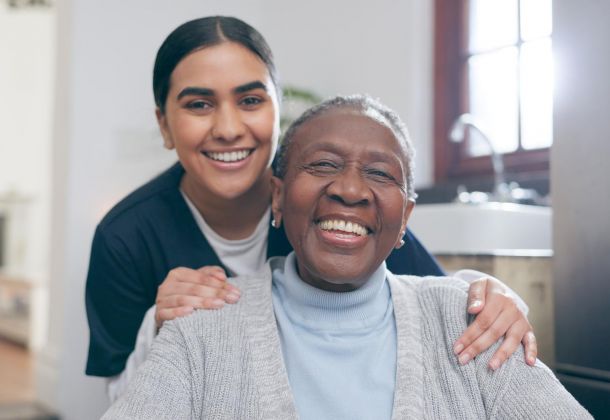 Smiling elderly woman sits while a younger woman stands behind her, both looking happy indoors.