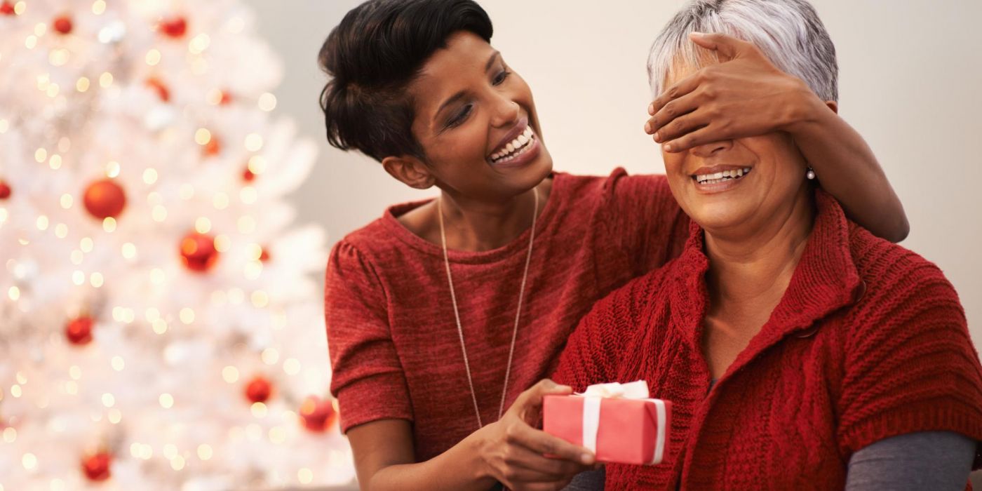 Young woman surprises smiling older woman with a gift in front of a decorated Christmas tree.