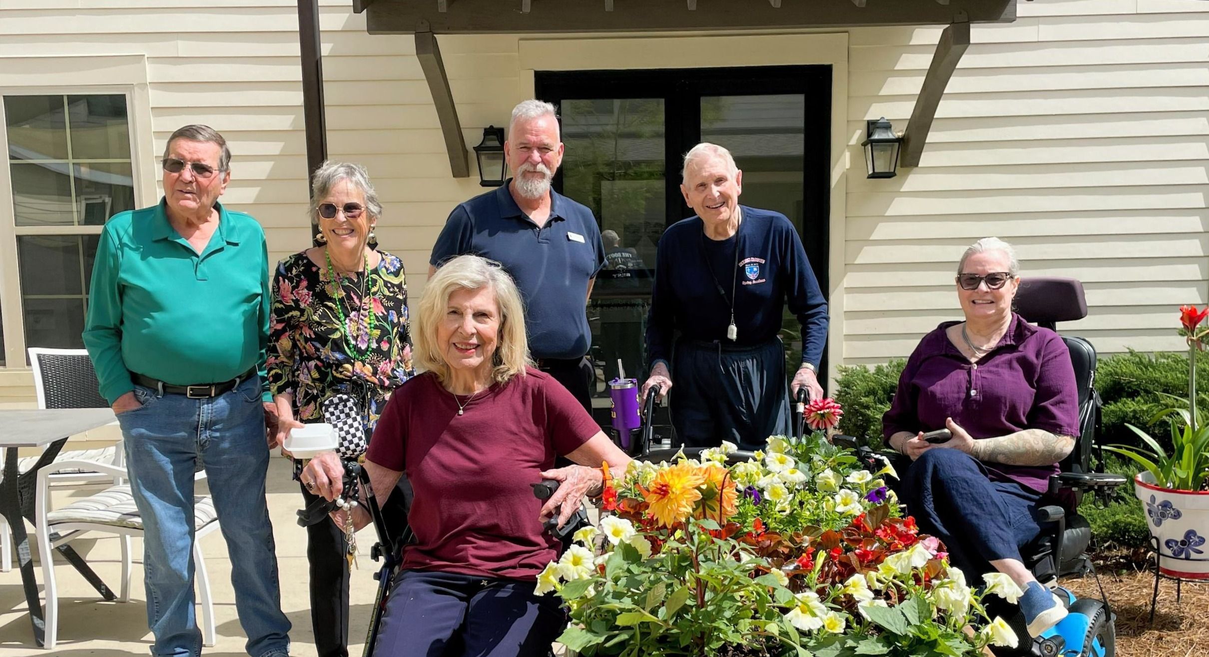 Six smiling older adults, some seated and some standing, pose outside by a colorful flower bed on a sunny day.