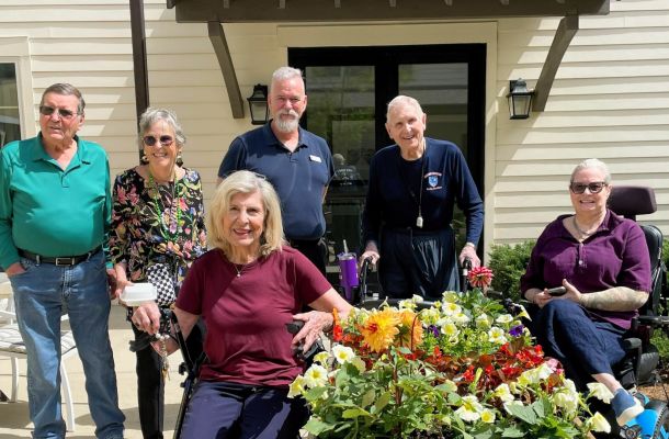 Six smiling older adults, some seated and some standing, pose outside by a colorful flower bed on a sunny day.
