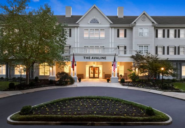 A large white senior living community named The Avaline with lit windows, two flags, and a landscaped driveway at dusk.