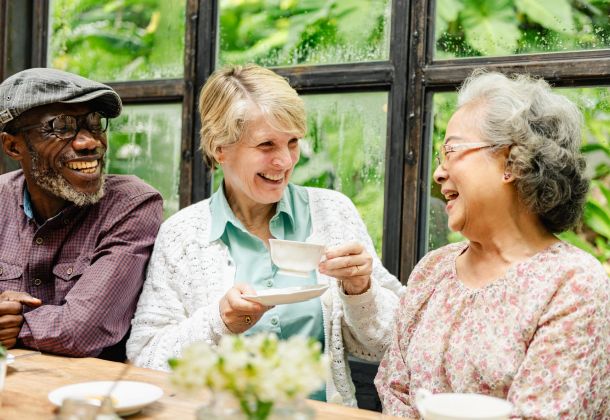 Three older adults laughing and enjoying tea together by a window with greenery outside.