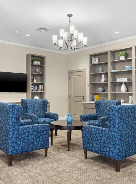 Room with blue patterned armchairs, round table, built-in shelves, and a wall-mounted TV under a chandelier.