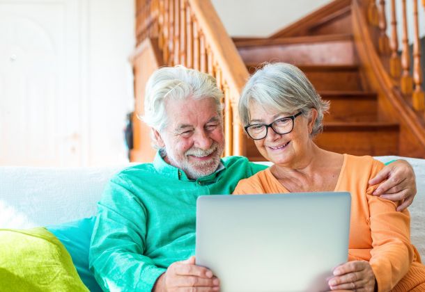 Smiling older couple sitting on a couch, looking at a laptop together in a cozy, sunlit living room with wooden stairs in the background.