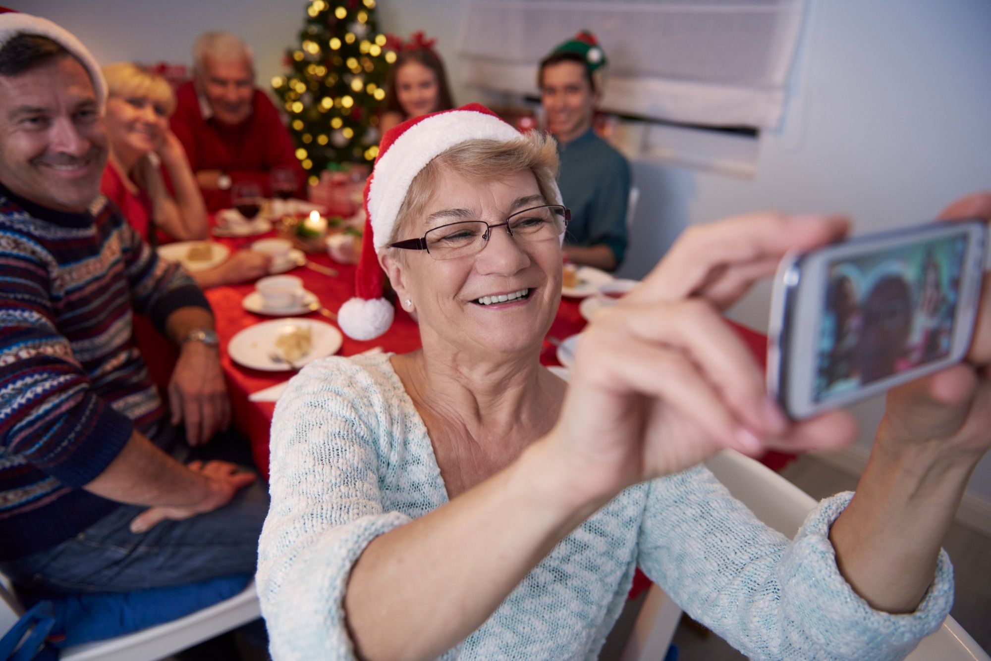 Smiling elderly woman in Santa hat taking a selfie with family at a festive table near a Christmas tree.