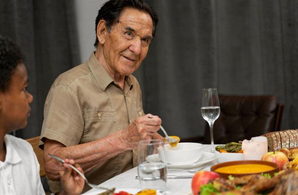 An elderly man eats soup at a dinner table, sitting beside a child, with food and drinks in front of them.