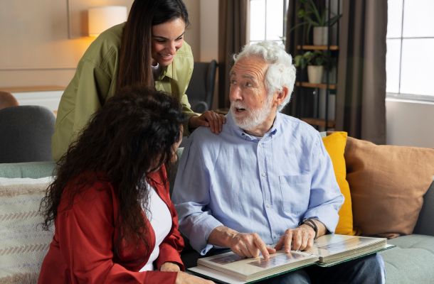 Older man showing a photo album to two women on a couch, all smiling and engaged in conversation.