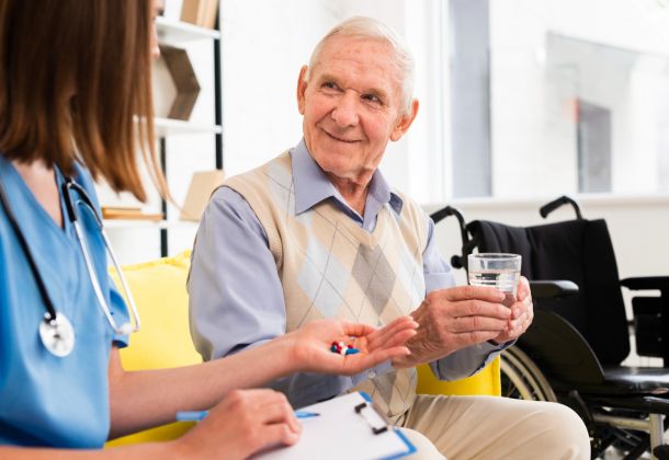 Elderly man holding a glass of water, receiving pills from a nurse, with a wheelchair in the background.