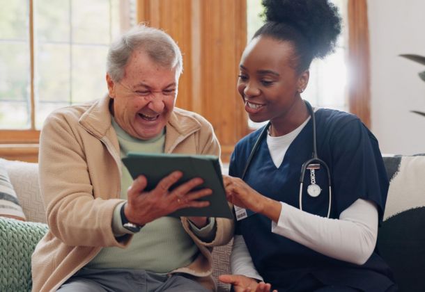 A nurse and an older man smile and look at a tablet together while sitting on a couch.