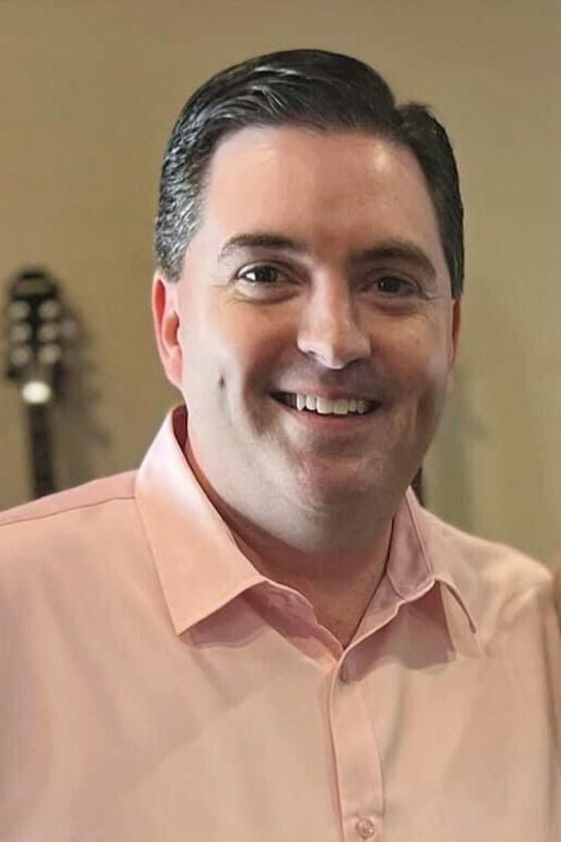 A smiling man in a light pink shirt stands indoors with guitars blurred in the background.