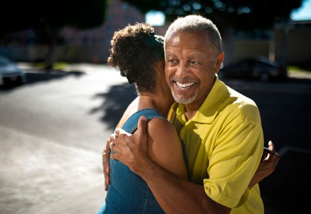 An older man in a yellow shirt smiles while hugging a woman outdoors on a sunny day.