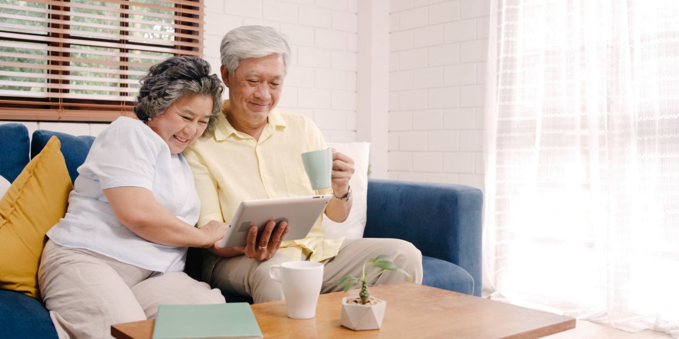 Smiling older couple sits on a couch, looking at a tablet together, with coffee mugs on a wooden table.
