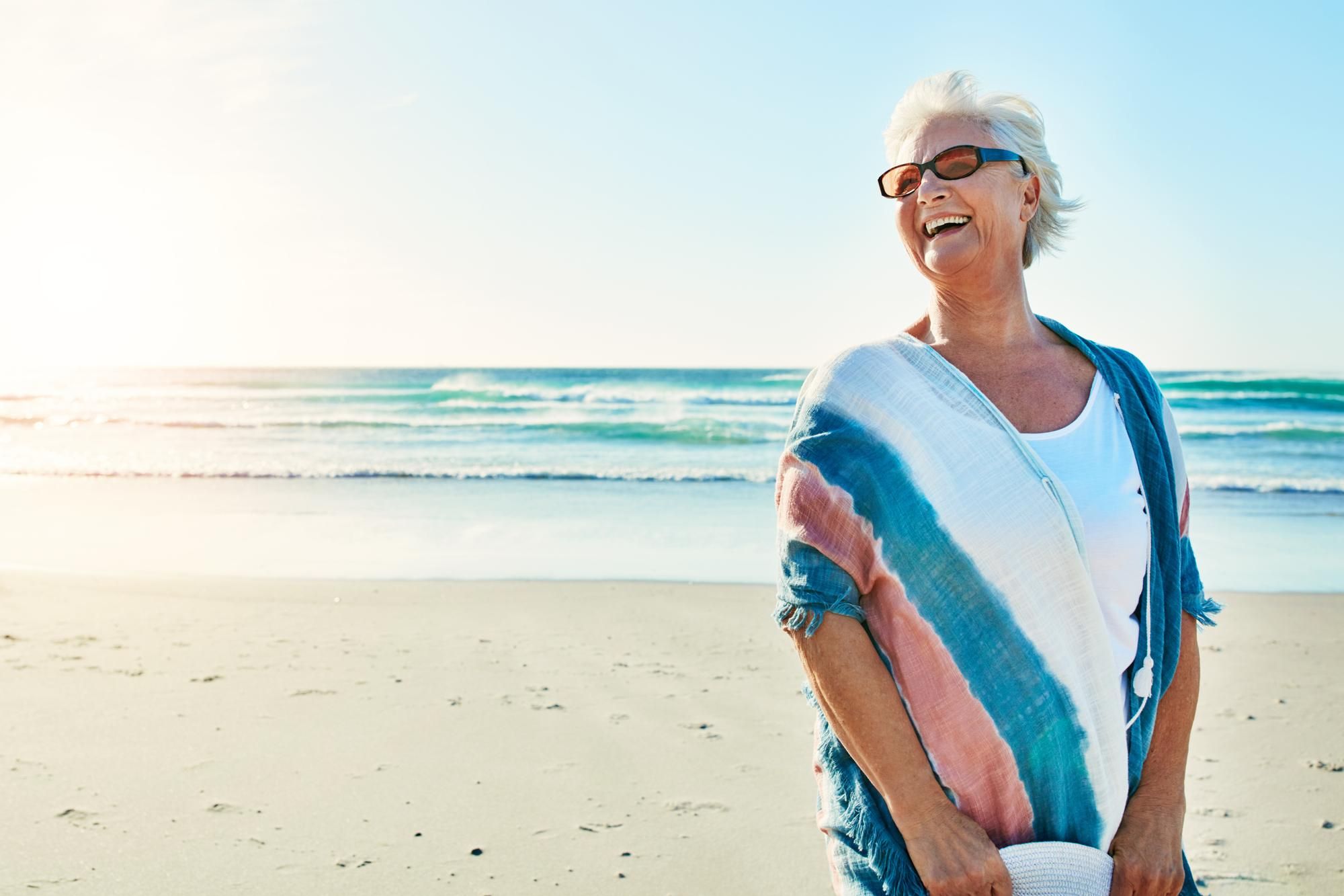 Smiling older woman in sunglasses stands on a sunny beach with waves in the background.
