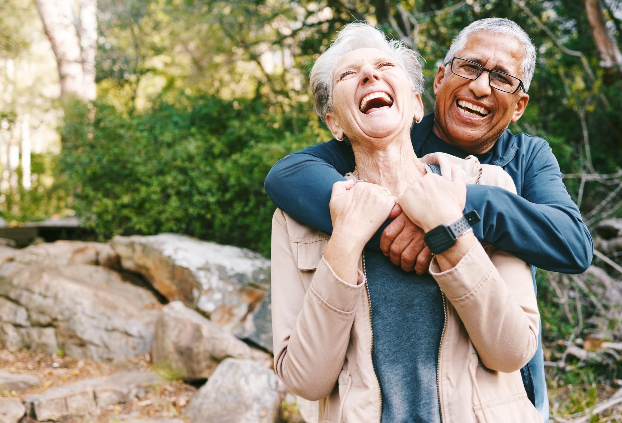 An older couple laughs joyfully as they hug each other outdoors in a wooded park.