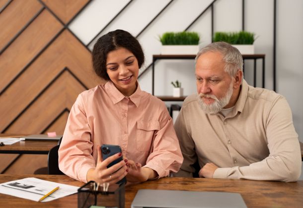 A young woman shows something on her phone to an older man as they sit together at a desk in a modern office.