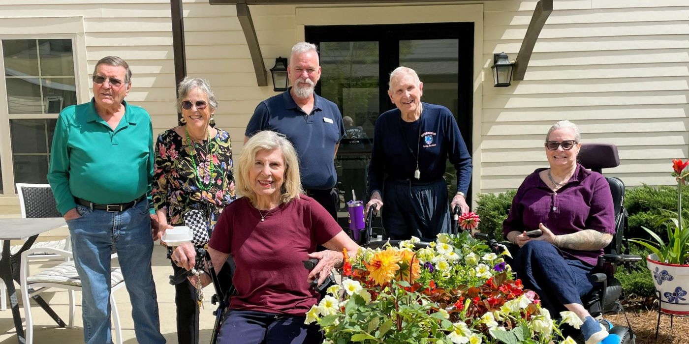 Six smiling older adults, some seated and some standing, pose outside by a colorful flower bed on a sunny day.