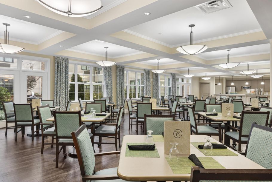 Bright dining room with empty tables, green chairs, and modern lighting. Large windows with patterned curtains.