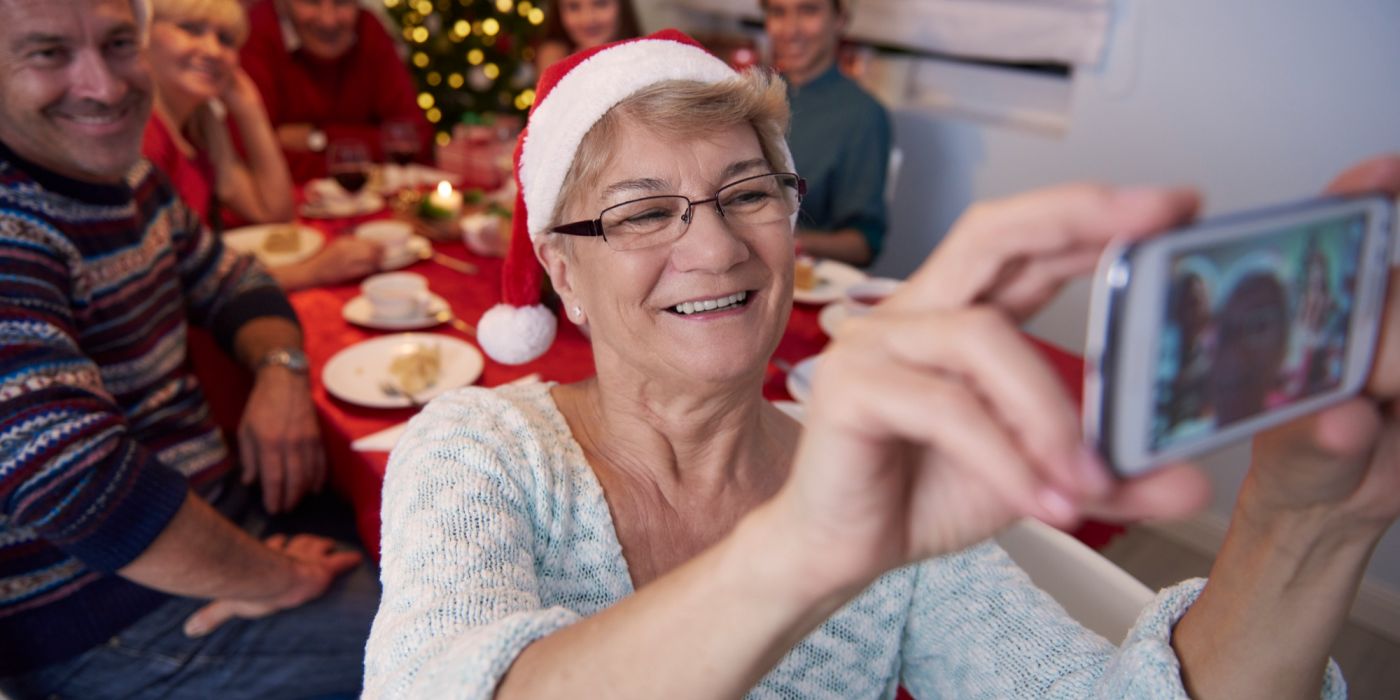 Smiling elderly woman in Santa hat taking a selfie with family at a festive table near a Christmas tree.