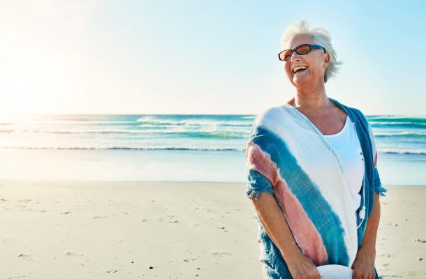 Smiling older woman in sunglasses stands on a sunny beach with waves in the background.