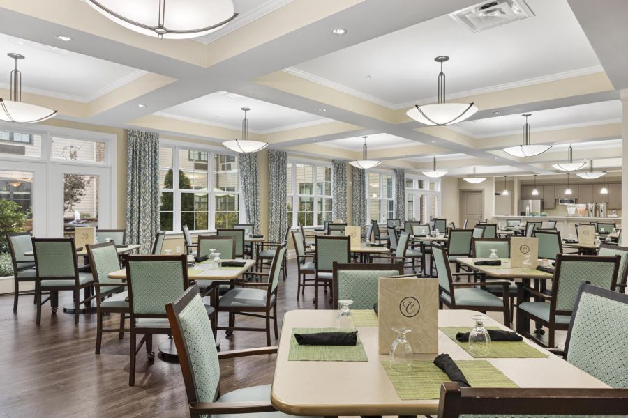 Bright dining room with empty tables, green chairs, and modern lighting. Large windows with patterned curtains.