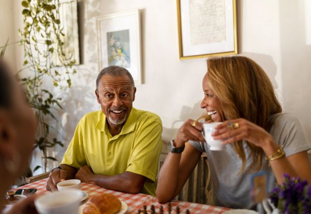 Two people sit at a table, smiling and drinking coffee together in a warmly lit room with framed art on the wall.