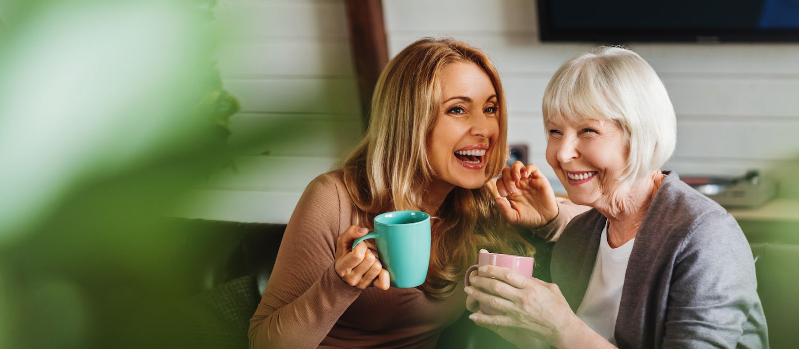 Happy senior mother with adult daughter sitting on couch and holding cups with coffee or tea at home. Togetherness concept