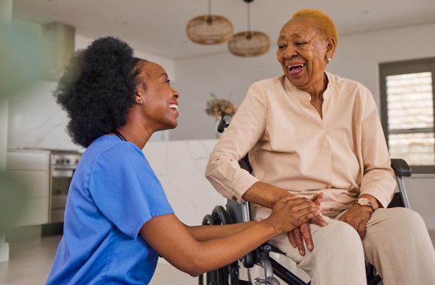 Nurse holding hands and smiling with an older woman in a wheelchair inside a bright home.