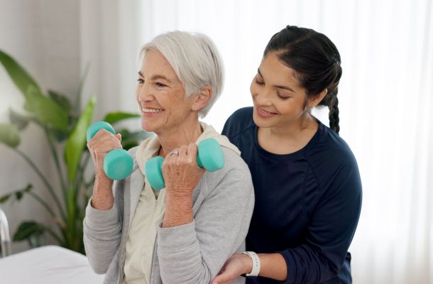 Elderly woman lifts dumbbells with assistance from a caregiver in a bright room.