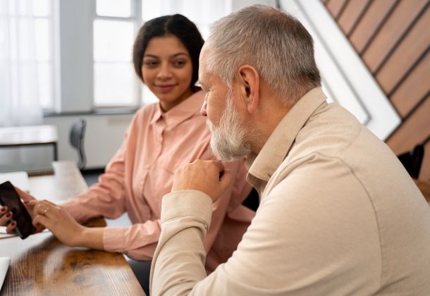 A young woman shows something on her phone to an older man as they sit together at a desk, talking.