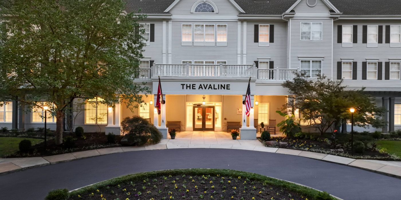 A large white senior living community named The Avaline with lit windows, two flags, and a landscaped driveway at dusk.