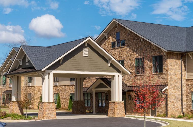 Brickmont at Johns Creek with stone facade, dark roof, covered entrance, and a small tree in front. Blue sky with clouds.