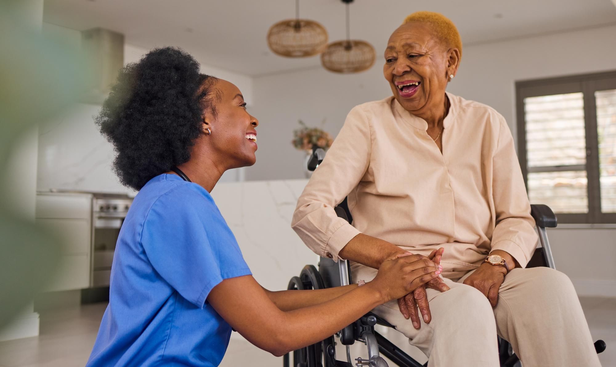 Nurse holding hands and smiling with an older woman in a wheelchair inside a bright home.
