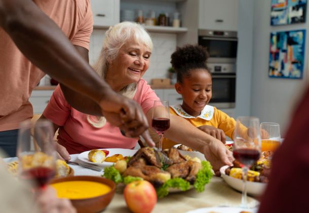 Smiling family sharing a meal at a table, with roast chicken, wine, and various dishes in a cozy kitchen.
