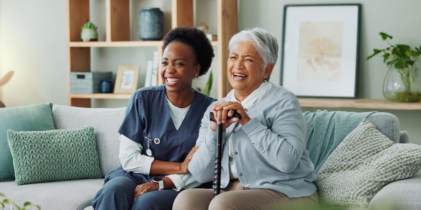 A caregiver and an older woman sitting on a sofa, both laughing.