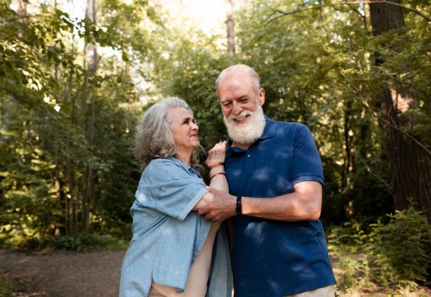 Smiling older couple standing together in a sunlit forest, enjoying each other's company.