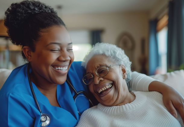 Nurse in blue scrubs with stethoscope hugs and laughs with an elderly woman wearing glasses and a white sweater.