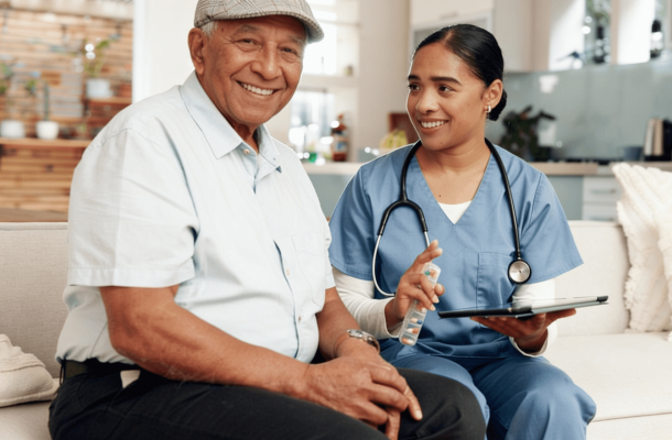 An elderly man smiles while sitting next to a nurse with a stethoscope and tablet in a bright living room.