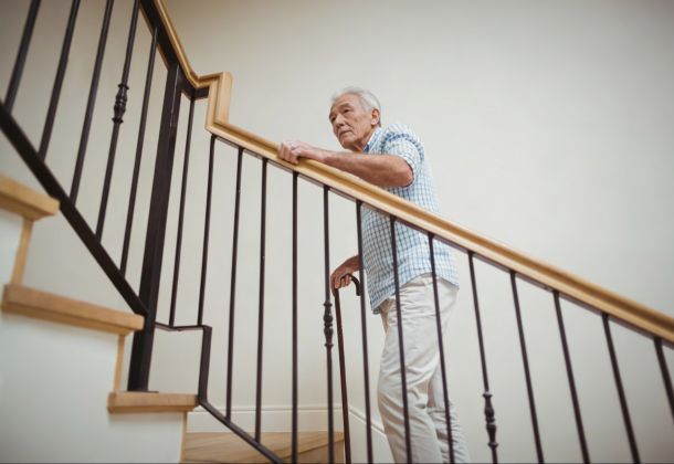 Older man in a checkered shirt holding a railing while walking up a staircase indoors.