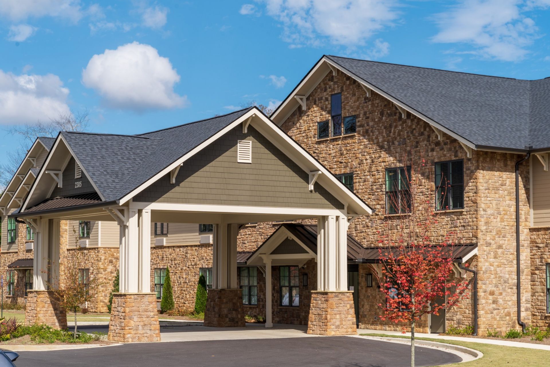 Brickmont at Johns Creek with stone facade, dark roof, covered entrance, and a small tree in front. Blue sky with clouds.